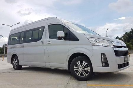 Silver Toyota Hiace minibus parked on a paved lot with trees and a cloudy sky behind it